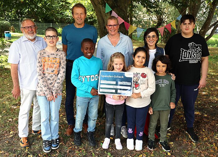 Vier Erwachsene und sechs Kinder mit einem Spendenscheck unter einem Baum mit Wimpelkette.