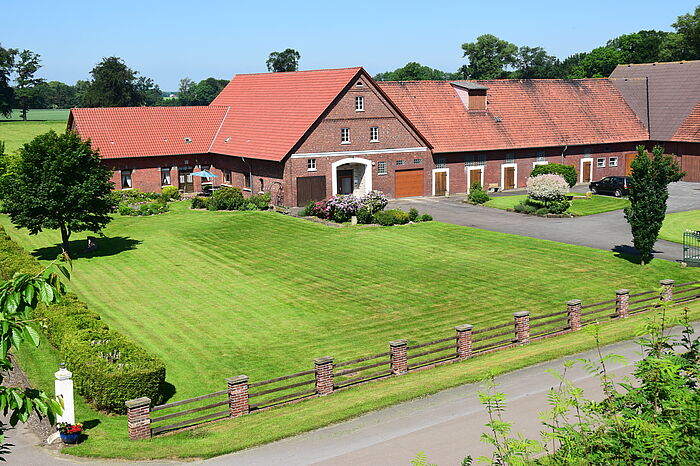 Rotes Bauernhof Haus mit Anbauten. Davor eine grüne Wiese, eingezäunt mit einem Zaun und an der linken HAnd mit einer Hecke