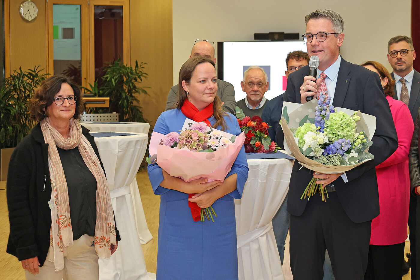 Zwei Frauen und ein Mann. Die Frau in der Mitte und der Mann halten Blumensträuße in der Hand.