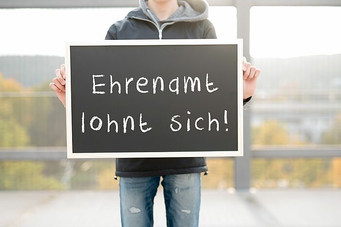 abgebildet ist eine Person, die eine Tafel vor sich hält, auf der mit Kreide steht: Ehrenamt lohnt sich!