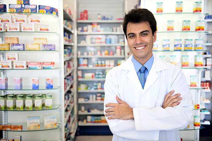 Ein Mann in weißem Kittel steht lächelnd mit verschränkten Armen vor mehreren mit Arzneimitteln gefüllten Regalen. 