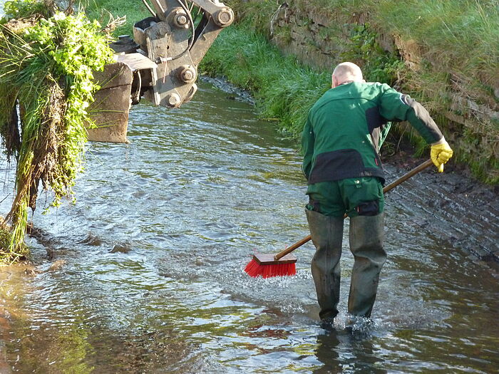 Ein Mann der mit einem Besen in einem Fluß steht und die Wasseroberfläche kehrt. Links ist ein Teil einer Baggerschaufel zu sehen.