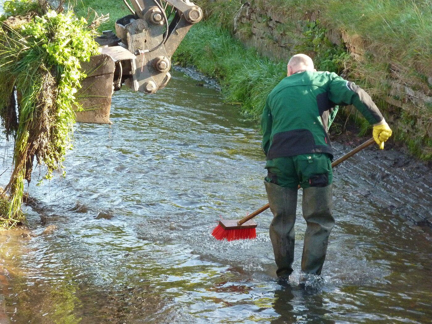 Ein Mann der mit einem Besen in einem Fluß steht und die Wasseroberfläche kehrt. Links ist ein Teil einer Baggerschaufel zu sehen.