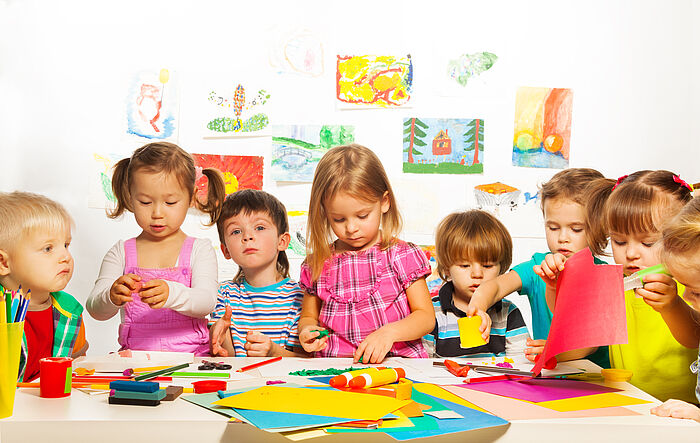 Acht Kindergarten-Kinder stehen an einem Tisch. Sie basteln und malen gemeinsam. Im Hintergrund hängen selbstgemalte Wachsbilder.
