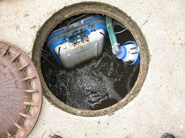 Draufsicht auf offenen Schacht von einer Kleinkläranlage, rechts zu sehen ist ein Probenahmebehälter, verbunden mit einer blau-weißen Kiste, die in das braune Abwasser Luft pumpt