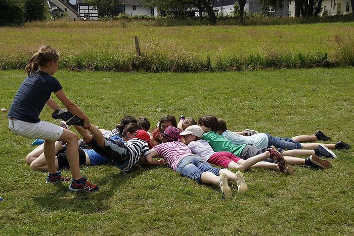 10 Kinder liegen auf dem Bauch im Kreis, wobei ein Mädchen ein Kind an den Beinen aus der Gruppe heraus zieht