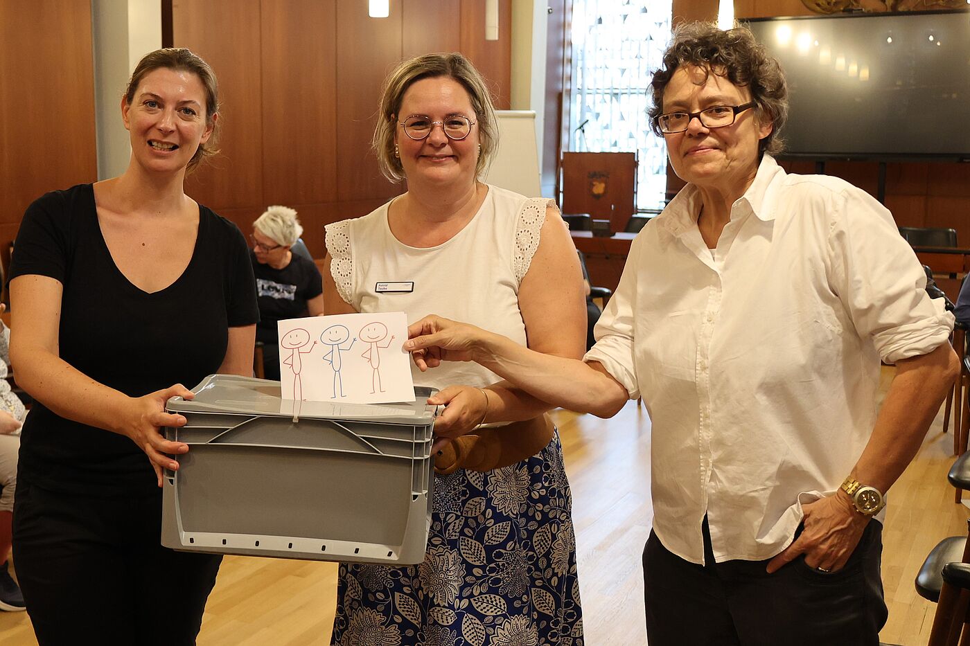 Drei Frauen halten einen Zettel mit Strichmännchen hoch.