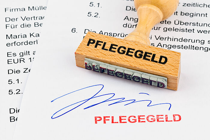 Zu sehen ist die untere Hälfte zweier bedruckter Blätter. Auf dem oberen Blatt liegt ein Holzstempel mit der Aufschrift "Pflegegeld". Dieser Stempel ist am unteren Rand des Bildes mit roter Tinte gesetzt worden. Darüber ist eine Unterschirft in blauer Tinte zu sehen.