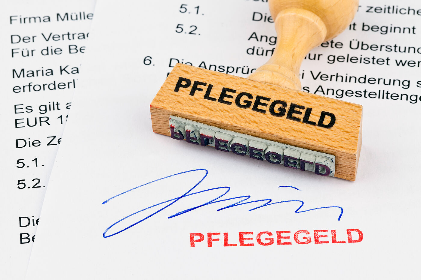 Zu sehen ist die untere Hälfte zweier bedruckter Blätter. Auf dem oberen Blatt liegt ein Holzstempel mit der Aufschrift "Pflegegeld". Dieser Stempel ist am unteren Rand des Bildes mit roter Tinte gesetzt worden. Darüber ist eine Unterschirft in blauer Tinte zu sehen.