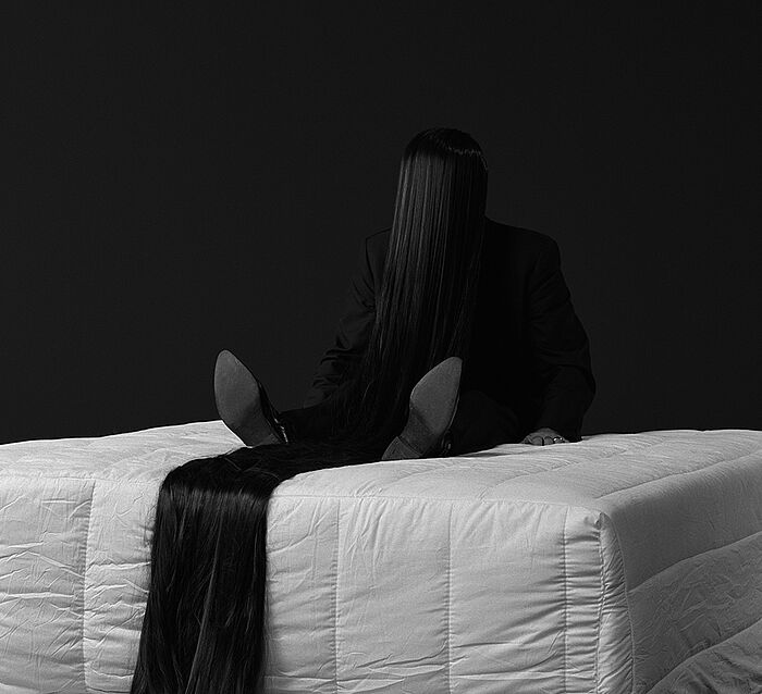 Person auf Bett sitzend, Gesicht durch überlange Haare verdeckt.