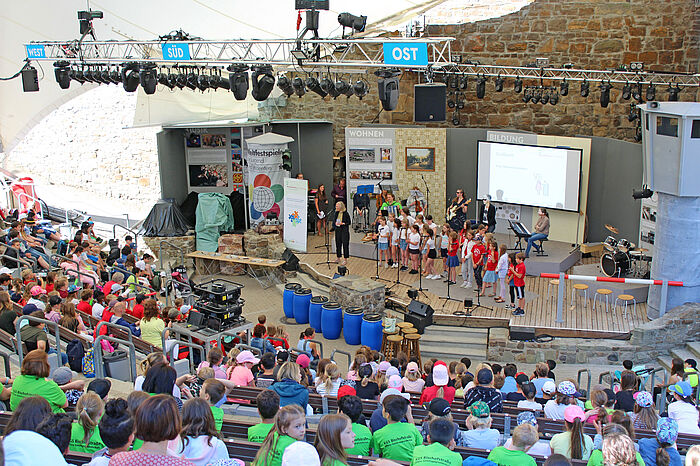Foto von einem Fest der musikalischen Grundschulen: Auf der Burg Wilhelmstein sitzen viele Kinder im Publikum und auch auf der Bühne treten Kinder auf