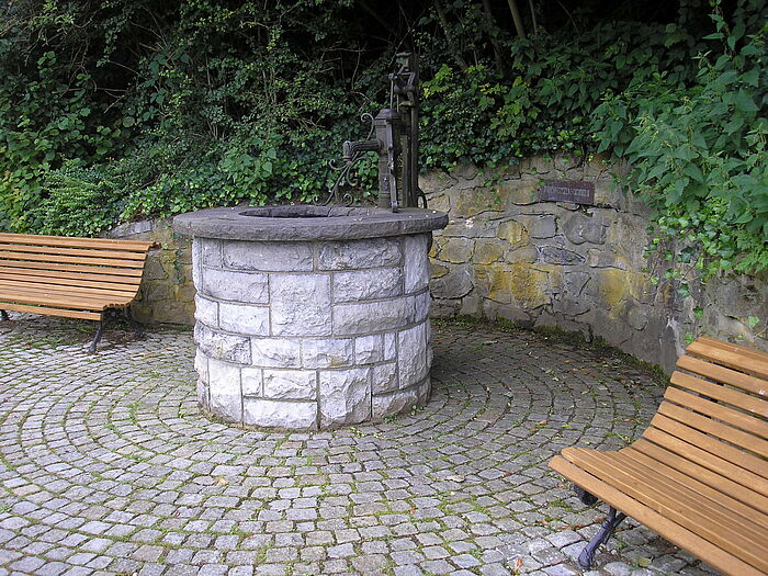 Ein runter Steinbrunnen mit alter Wasserpumpe auf einem Platz der mit Pflastersteinen rund um den Brunnen gepflastert ist. Zwei Holzsitzbänken stehen jeweils neben dem Brunnen. Im Hintergrund sind Pflanzen.