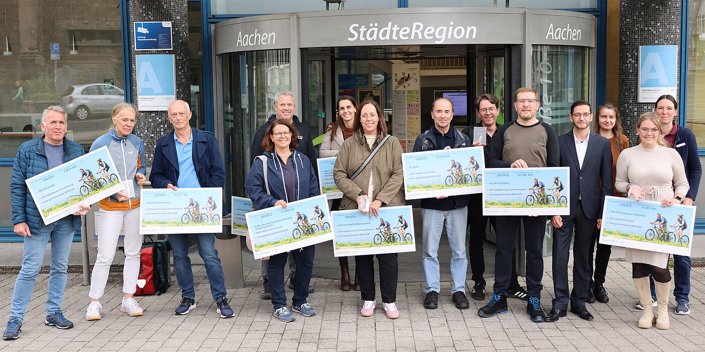 Menschen mit großen Schecks in der Hand vor einem Gebäude.