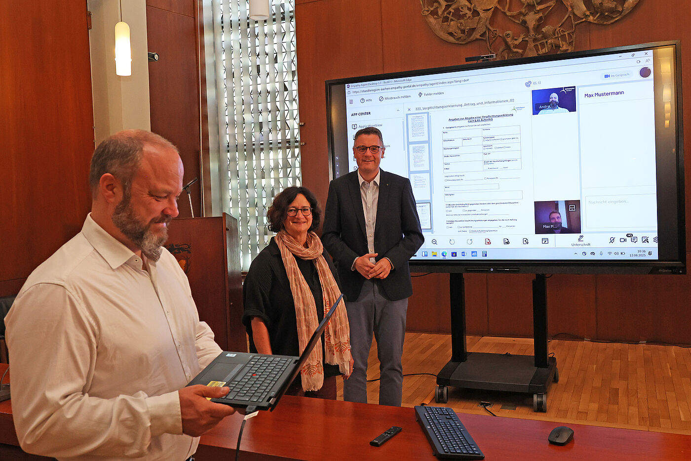 Ein Mann hält einen Laptop in der Hand. Im Hintergrund stehen eine Frau und ein Mann vor einem großen Bildschirm auf dem das Gesicht des Mannes mit dem Laptop zu sehen ist.