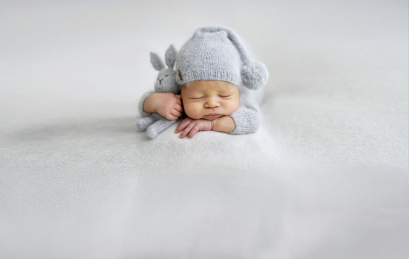 Ein Baby mit grauer Mütze schläft mit einen kleinen grauen Stoffhasen im Arm