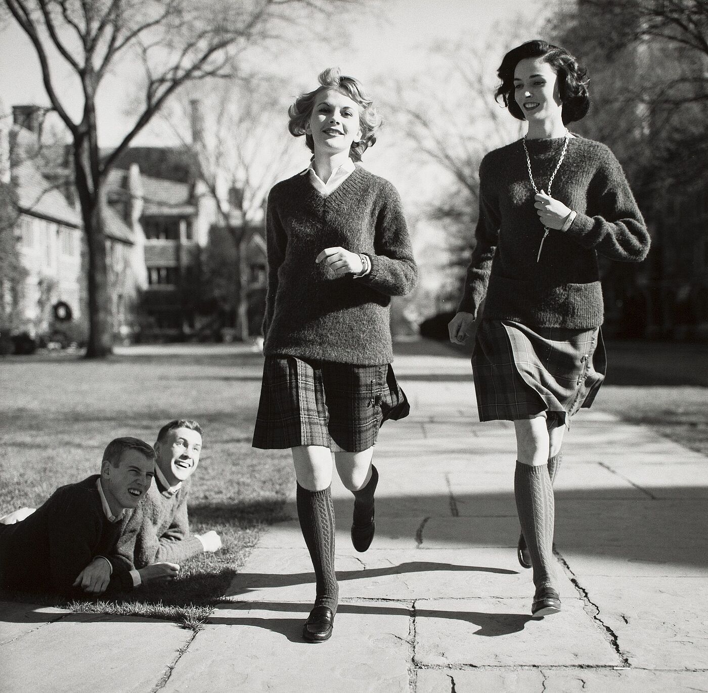 Zwei Frauen laufen über einen Gehweg und werden dabei von zwei Männern beobachtet