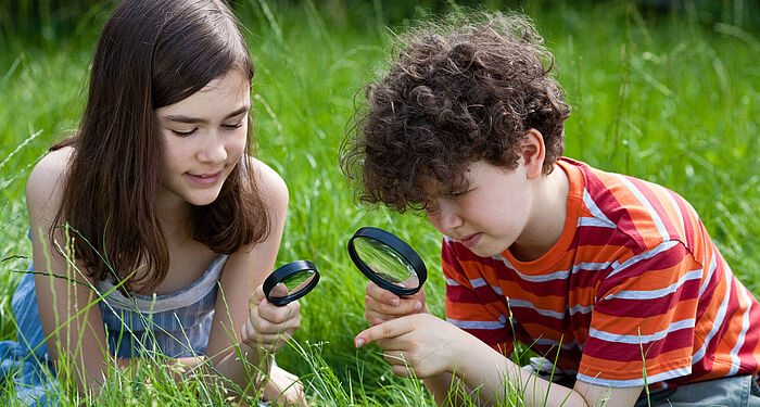 zwei Kinder sitzen auf einer Wiese und untersuchen Grashalme mit Lupen