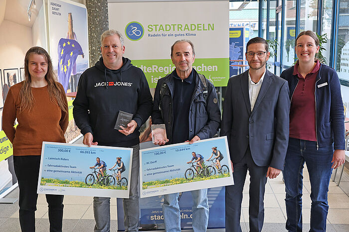 Vier Männer und zwei Frauen vor einem Plakat mit großen Schecks in der Hand.