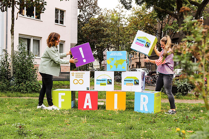 Zwei Frauen stapeln auf einer Wiese große bunte Würfel aufeinander, die mit Begriffen zum Thema Fairtrade beschriftet sind.