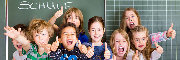 Fröhliche Grundschulkinder vor einer Tafel. Auf der Tafel steht "Schule".