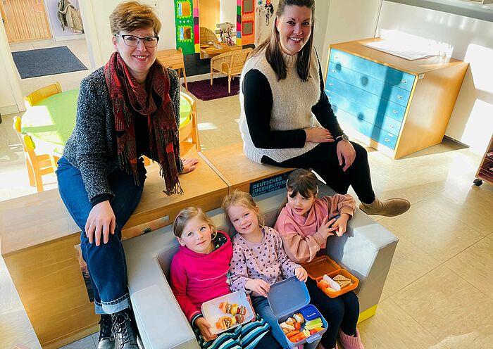 drei Kinder in einer Kita sitzen auf einem Sofa. Neben ihnen sitzen eine Erzieherin und eine Beraterin aus dem Gesundheitsamt