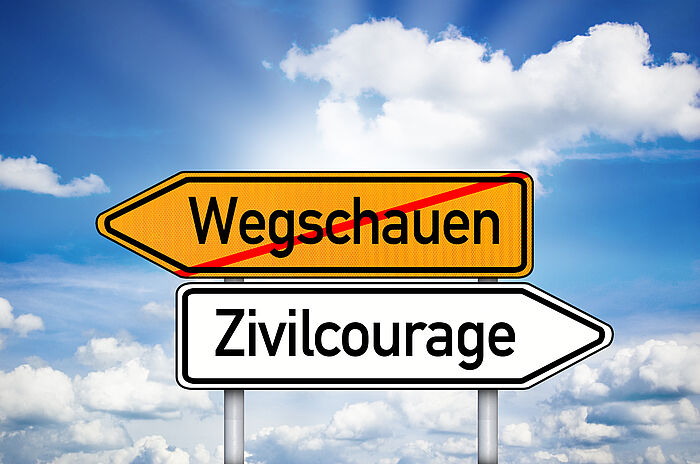 Man sieht vor einem blauen Himmelhintergrund mit weißen Wolken zwei Straßenschilder,die in zwei verschiedene Richtungen zeigen. Das gelbe Schild zeigt nach links und hat die mit rot durchgestrichene Aufschrift  "Wegschauen".  Das weiße Schild zeigt nach rechts und hat die Aufschrift "Zivilcourage".