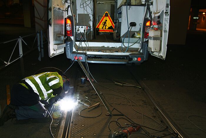 Zu sehen ist ein Mann mit Sicherheitsweste, der in der Nacht auf den Boden Metallschienen schweißt. Sein Fahrzeug mit den Gerätschäften steht im Hintergrund.