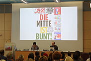 "Die Mitte ist bunt" steht zu lesen im Hintergrund zweier Referenten auf der Bühne des Fachtages.