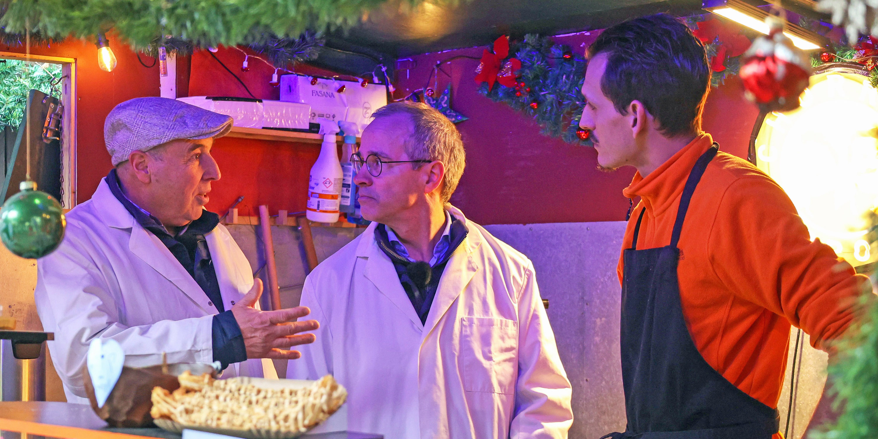 Zwei Männer mit Kittel und einer mit Schürze in einem Weihnachtsmarktstand.