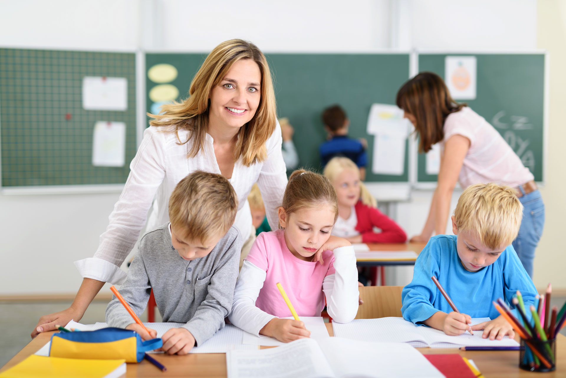 Lehrerin hinter Grundschüler_innen stehend, die mit Stiften schreiben 