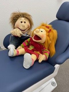 Eine Puppe sitzt mit geöffnetem Mund auf einem zahnärztlichen Untersuchungsstuhl und eine weitere Puppe ist der Zahnarzt