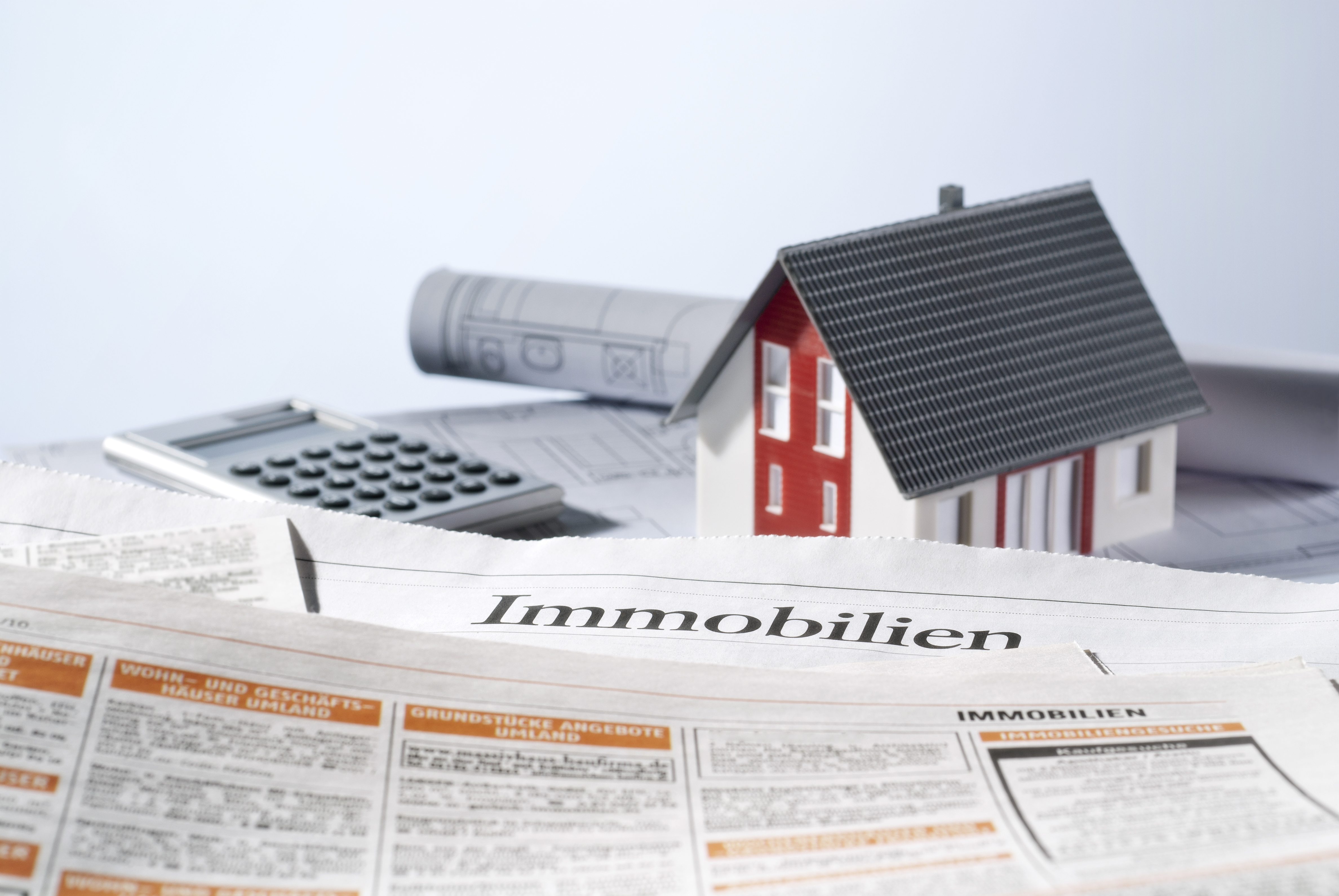 Tisch, auf dem der aufgeschlagene Teil einer Zeitung mit Immobilienteil, ein kleines Haus, Baupläne und ein Taschenrechner stehen