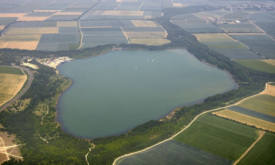 Luftbildaufnahme vom Blausteinsee. Zu sehen ist der See in seinem vollen Umfang, umgegeben von verschiedenen Feldern und grünen Baumstreifen