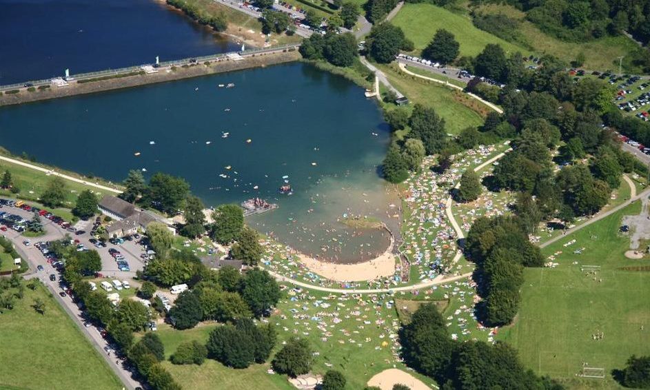 Luftbildaufnahme vom Eiserbachsee Rurberg, zu sehen ist der Badebereich mit der Staumauer, Menschen im Wasser und auf der Wiese liegend