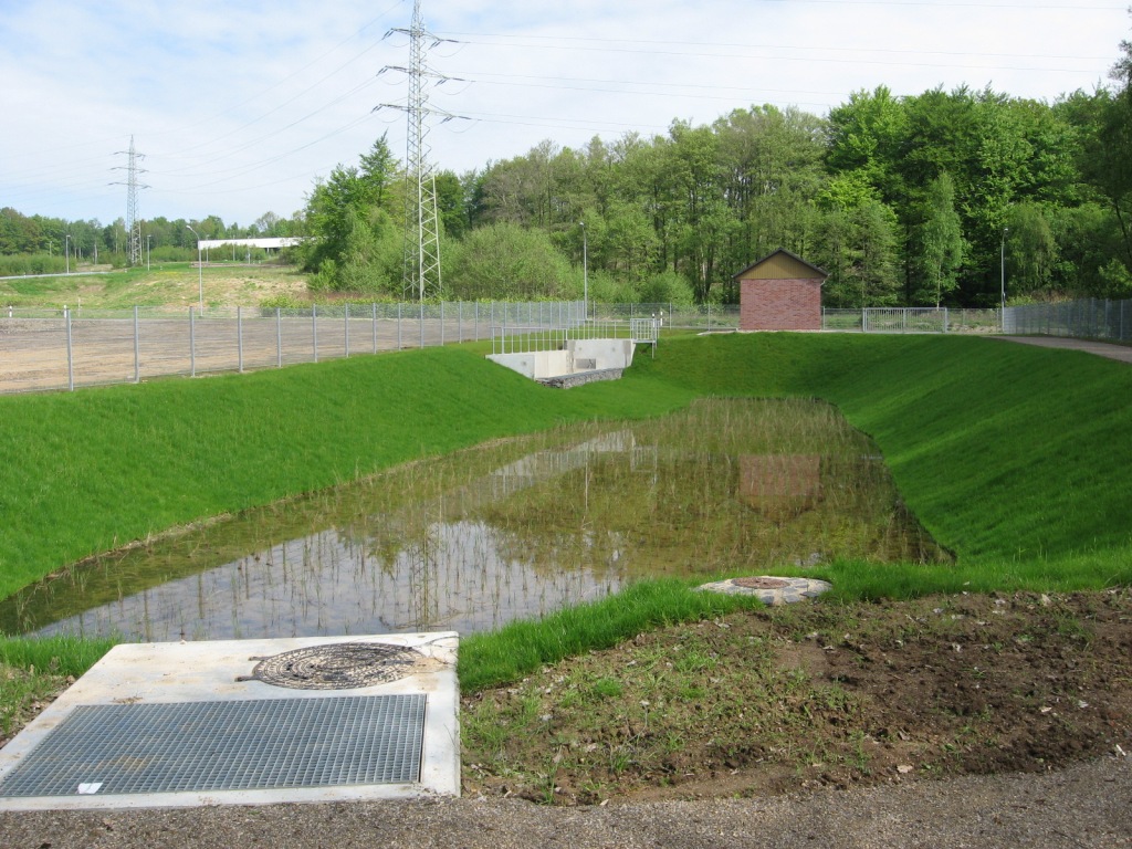 Eingezäunter Bereich, Bodenfilterbecken (Wasserloch) mit etwas Wasser gefüllt. Im Hinteren Bereich ist ein Kontrollhäuschen mit roten Ziegelstein, ohne Fenster, zu sehen. Hinter dem Zaun sind Bäume und Strommäste.