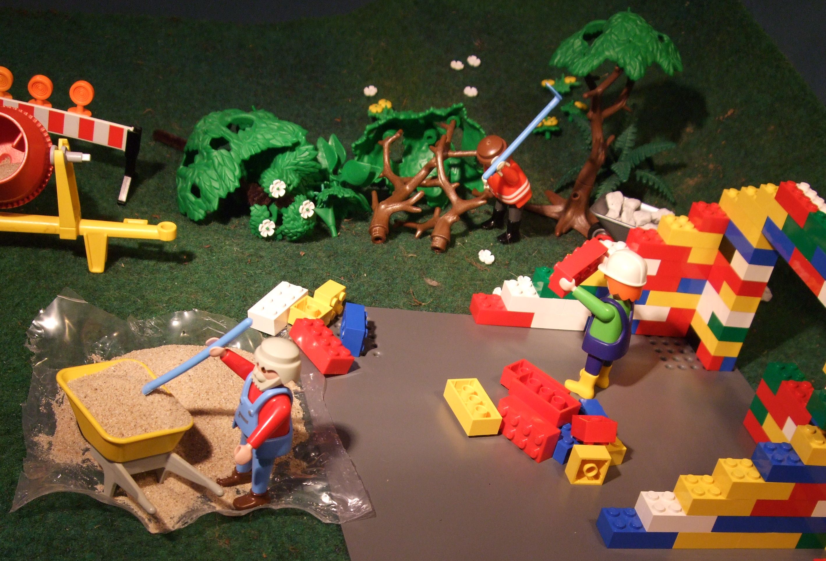 Mit Playmobilfiguren ist eine Baustelle nachgestellt worden. Zu sehen ist links ein Männchen mit Schubkarre, welches Sand schaufelt. Rechts gibt es ein angefangens Legohaus, wo ein Männchen einen Bauklotz in den Händen hält und im hingeren Teil zerkleinert ein Männchen liegende Bäume. Eine Betonmischmaschine ist hinten links auch noch zu sehen.