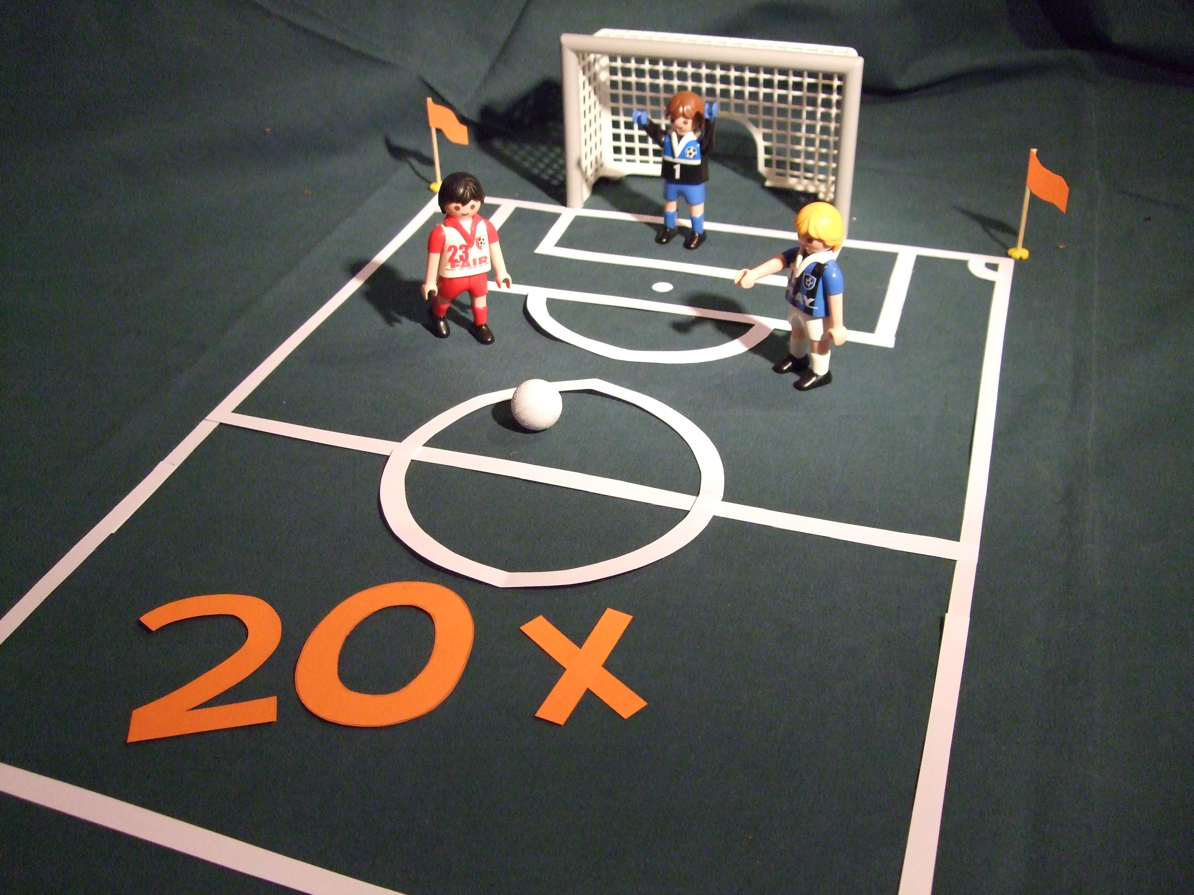 Mit Playmobile Figuren wurde ein Fußballspielfeld nachgestellt. Zu sehen ist das Spielfeld, ein Tor und 3 Playmobilfiguren. Im Spielfeld ist im vorderen Bereich eine 20 x in oranger Schrift aufgeklebt worden