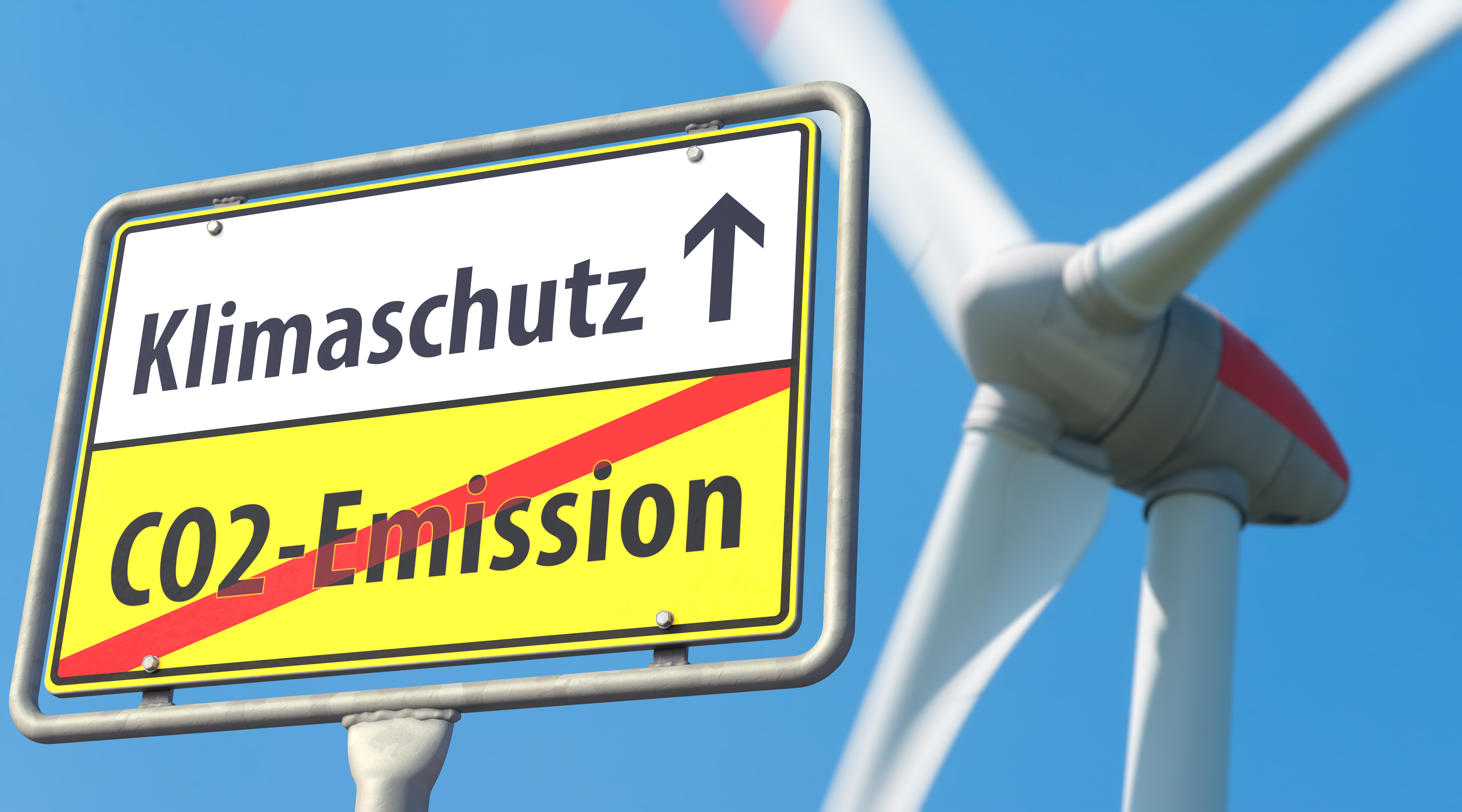 Ein Straßenschild mit dem Wort Klimaschutz und darunter durchgestrichen das Wort CO2 Emission, im Hintergrund einen Propeller vom Windkrafwerk