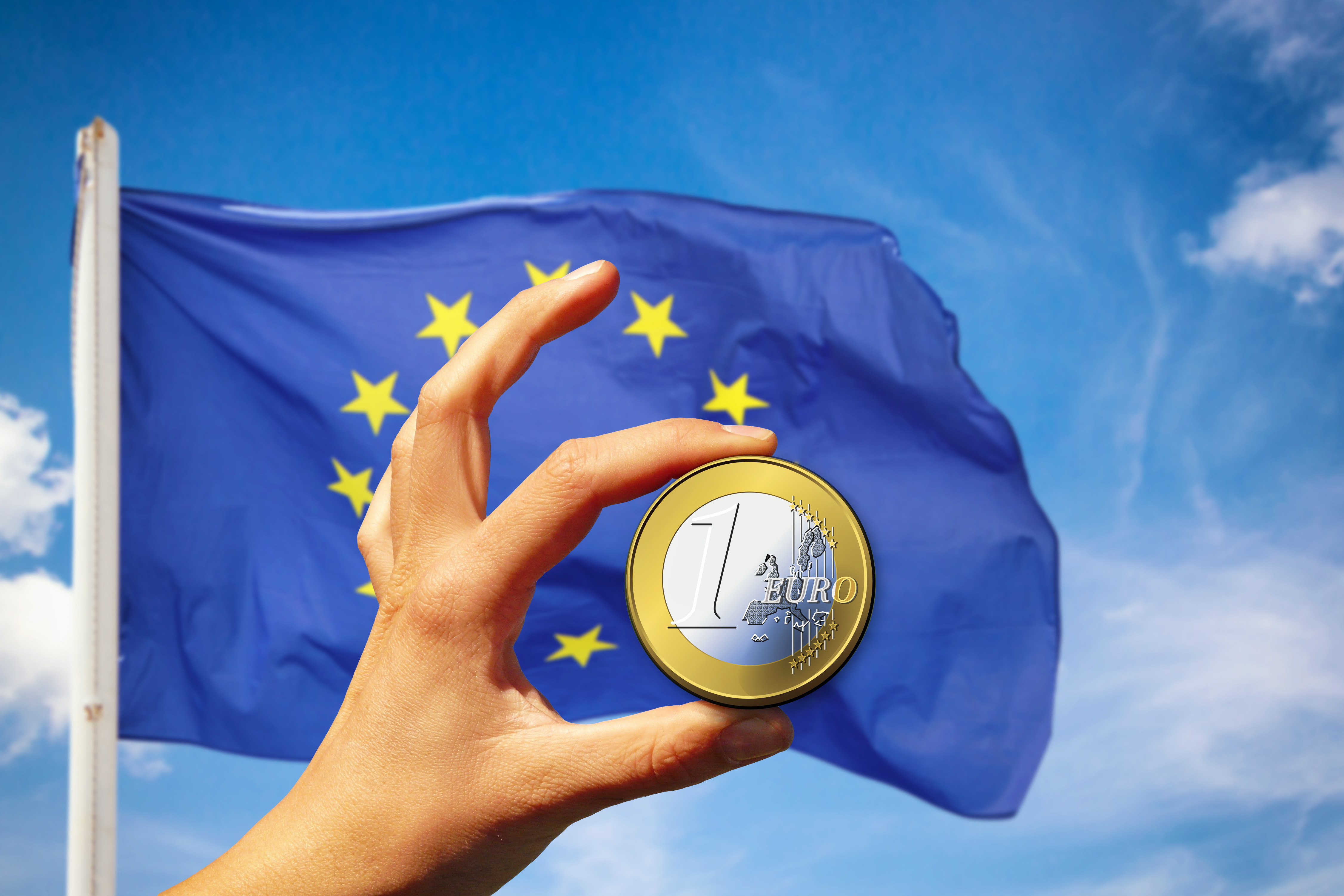 Eine Hand hält eine Euromünze vor einer europäischen Fahne