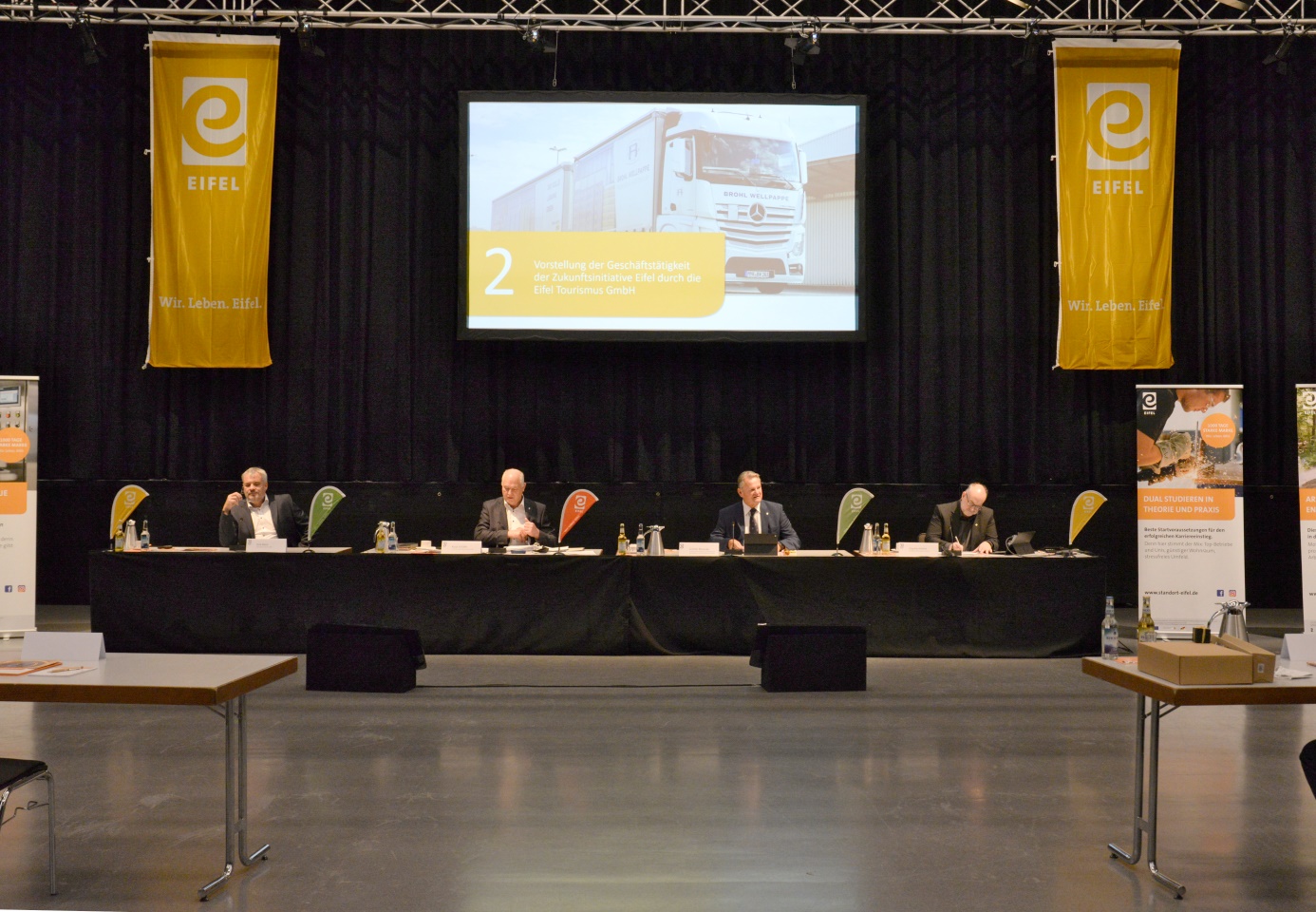 Leinwandpräsentation in einem großen Saal. darunter sitzen 4 Vortragende