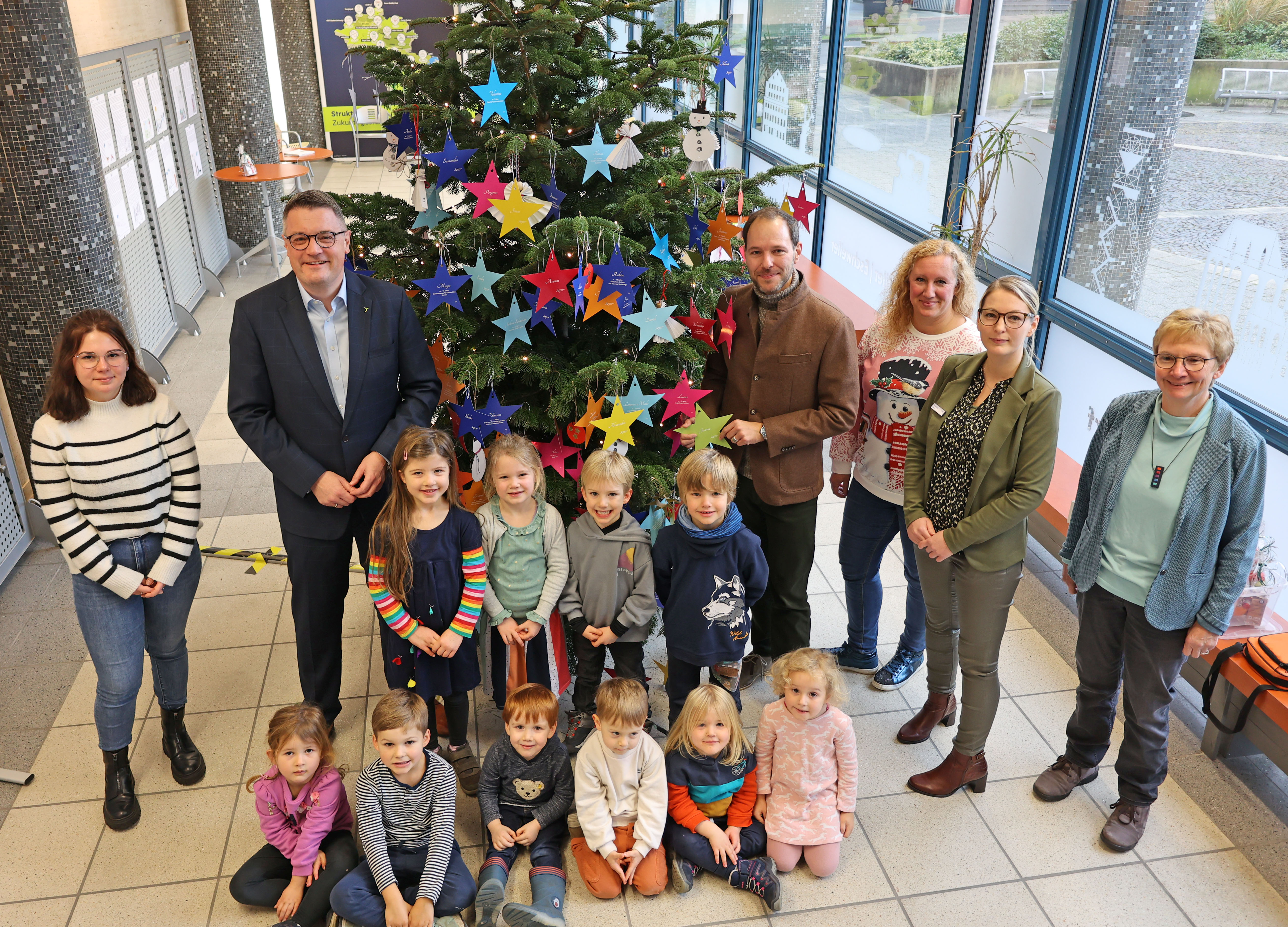 eine Gruppe Kita-Kinder steht mit Erzieherinnen und Städteregionsrat Tim Grüttemeier vor einem Weihnachtsbaum, der mit Wunschsternen geschmückt ist 
